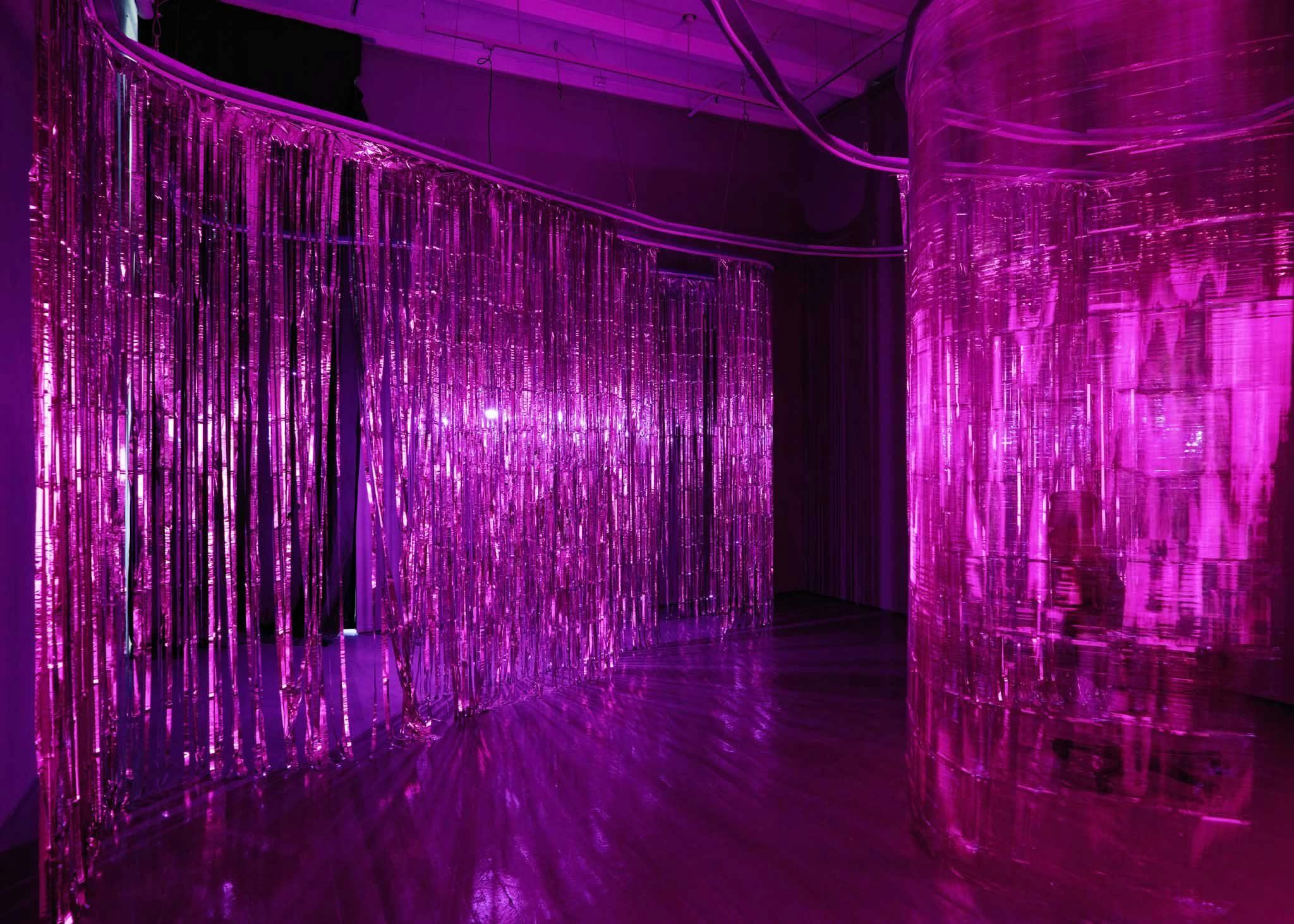 Foil curtains are caught in motion, highlighted by a fushcia light in a dark gallery