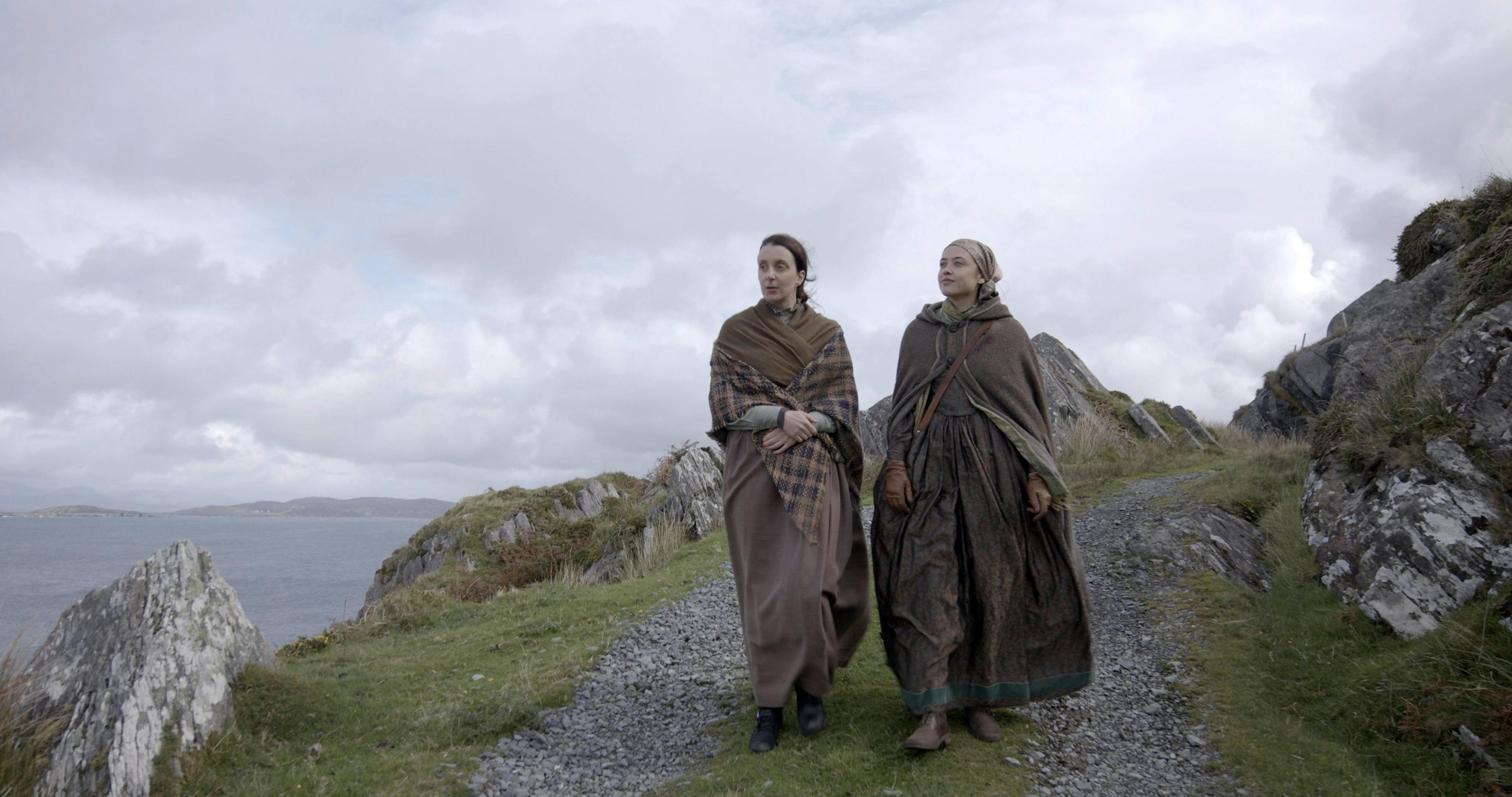 Two women in brown cloaks walk along a stone path by the sea.