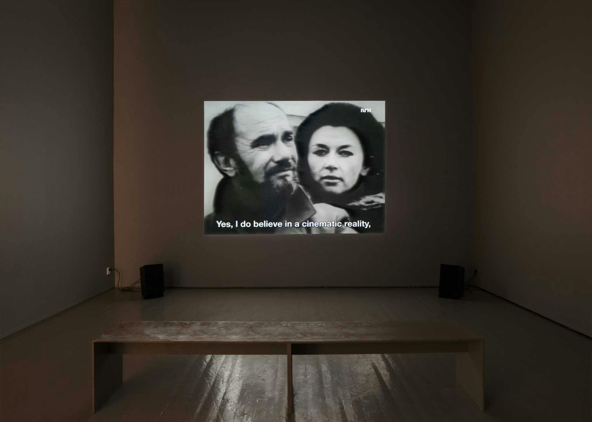 A bench sits in front of a projected image of a man and a woman in a grey gallery
