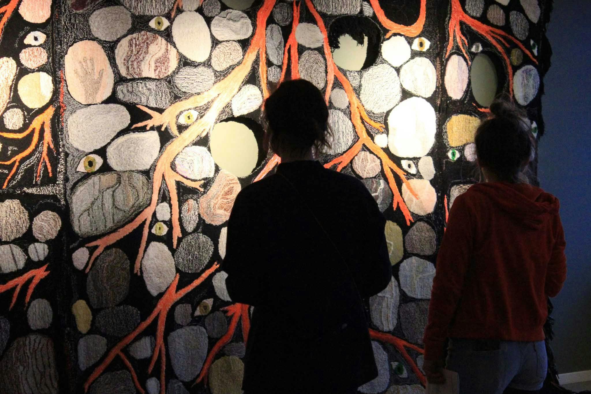 Two people stand in front of a large textile depicting roots and eyes
