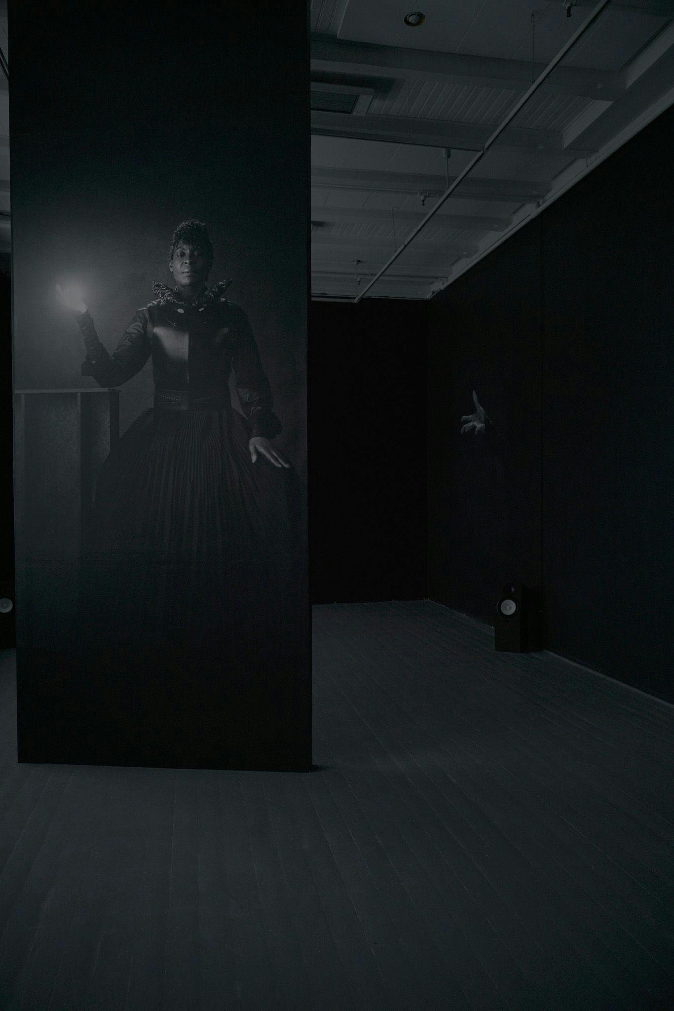 A woman in a black dress holding an orb of light is projected onto a black wall, in a black room.