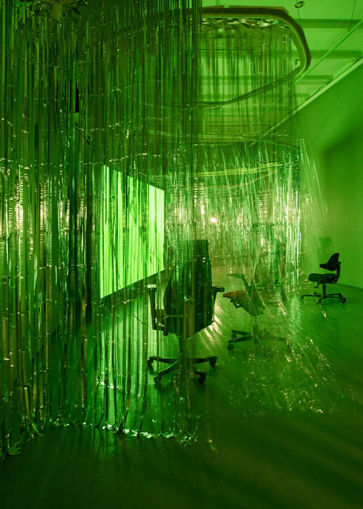 Office chairs are surrounded by foil curtains in a gallery space. Green light from a projection screen lights up the space.